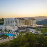 Modern resort hotel complex with a large outdoor pool, loungers, and landscaped grounds at sunset on a hillside.