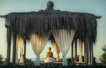 Person meditating cross-legged inside a thatch-roof gazebo with white drapes by the sea at sunset/sunrise.