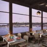 Sunset-lit waterfront restaurant with tables set for dining along large windows overlooking a calm bay and cityscape beyond.