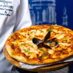 Pizza Italiante, Konya’da Açılıyor Chef in a white jacket presents a seafood pizza on a wooden board, topped with mussels and dill.