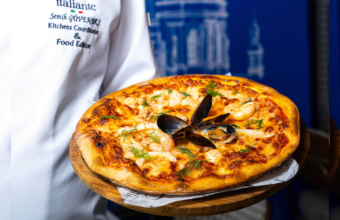 Chef in a white jacket presents a seafood pizza on a wooden board, topped with mussels and dill.