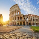 Sun setting behind the Colosseum in Rome, casting warm light across the cobblestone foreground.