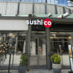 SushiCo storefront with glass doors and a round red logo, flanked by potted plants outside the entrance.
