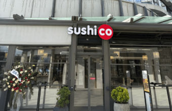 SushiCo storefront with glass doors and a round red logo, flanked by potted plants outside the entrance.