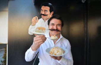 Smiling man in a white shirt holding plates of rice, with a cartoon chef cutout behind him on a dark wall.