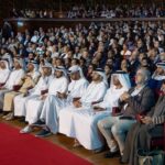Audience seated in a large conference hall, many in traditional Emirati attire, watching a talk with focus on the stage area on the left.