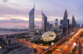 Aerial view of Dubai at dusk with futuristic skyscrapers and a large decorative ring sculpture near a busy highway.