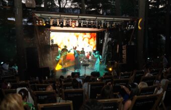 Outdoor night concert: a band performing on a lit stage with musicians, dancers, and a drum set, audience seated in rows watching.