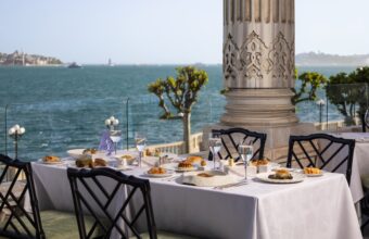 Outdoor seaside dining table set with white tablecloth, plates of pastries and bread, wine glasses, and a stone ornate column against the ocean backdrop.