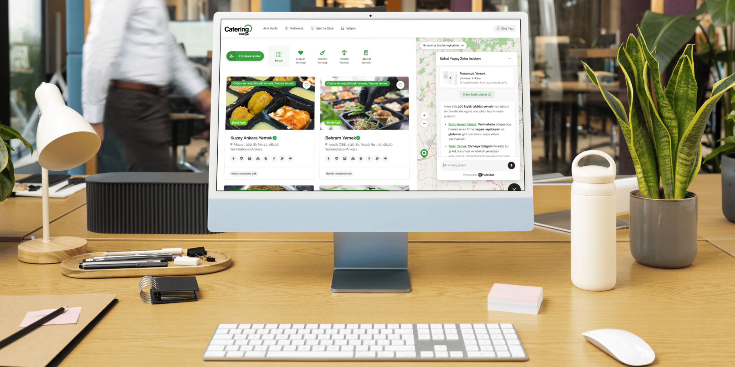 Desktop monitor on a wooden desk displaying a catering app with meal cards and a map panel on the right side.