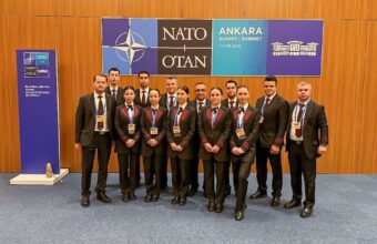 Group of well-dressed delegates posing for a photo at a NATO summit in Ankara, with badges visible and banners in the background.