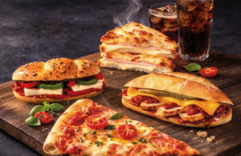 Assorted deli sandwiches and a cheese pizza slice on a wooden board with a glass of cola in the background.