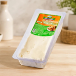 Tahsildaroğlu’ndan Listeria Açıklaması: İlgili Parti Piyasadan Çekildi Tilted package of white cheese on a wooden kitchen table, with a green-and-orange label reading its product name and a blurred milk bottle and fruit bowl in the background.