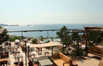 Terrace with tan-striped seating and wooden tables overlooking the sea, with a pool area in the foreground and distant islands on the horizon.