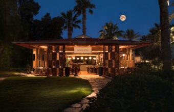 Open-air restaurant pavilion at night with warm lighting, outdoor tables, and palm trees under a dark blue sky with a full moon.