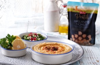 Hummus dip in a white bowl with paprika, lemon wedges and parsley on a silver tray, with a small salad and olive oil nearby.