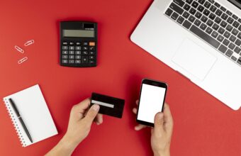 Two hands hold a black credit card and a smartphone on a red desk, with a calculator, notebook, and laptop nearby (payment setup).