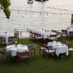 Outdoor seaside dining setup with round tables covered in white cloths on a green lawn by the water in the harbor area.