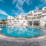 Outdoor hotel pool area with white, modern buildings and lounge chairs under a bright blue sky, during daytime.