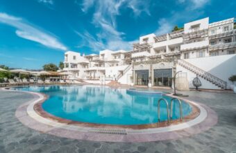 Outdoor hotel pool area with white, modern buildings and lounge chairs under a bright blue sky, during daytime.