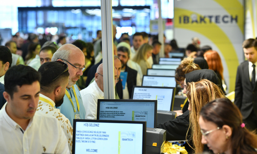 Crowd of attendees at a tech expo, standing at row of computer kiosks with staff helping visitors (IBAKTECH booth).