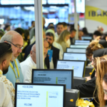 Crowd of attendees at a tech expo, standing at row of computer kiosks with staff helping visitors (IBAKTECH booth).