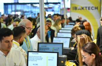 Crowd of attendees at a tech expo, standing at row of computer kiosks with staff helping visitors (IBAKTECH booth).