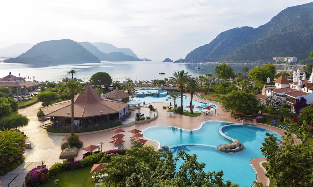 Overhead view of a tropical resort with winding blue pools, palm trees, thatched pavilions, and a calm bay with mountains in the distance.